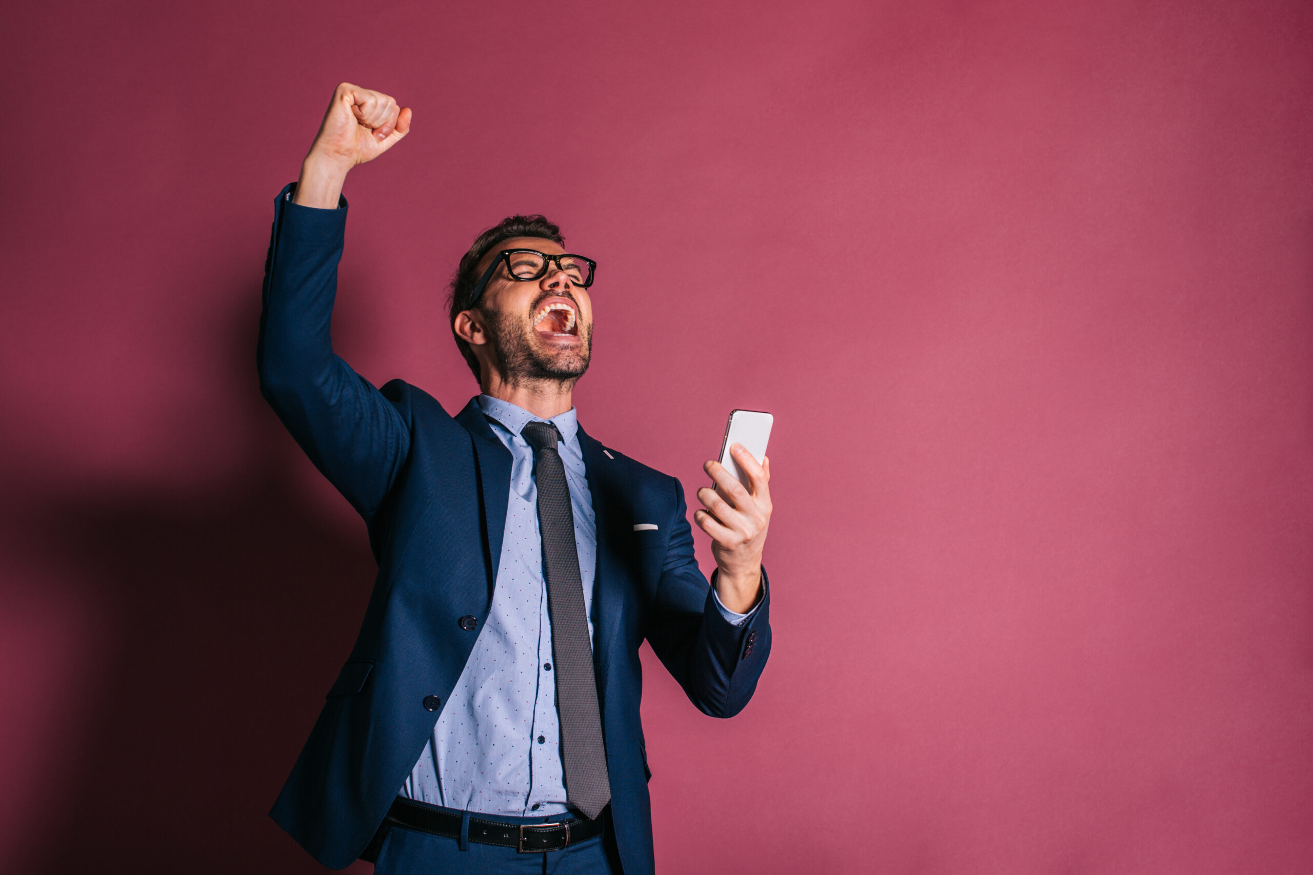 Homme d'affaire qui tient son téléphone cellulaire et qui lève le poings en l'air, tout en regardant au ciel, criant sa joie d'avoir conclue une vente grâce à C.A.I.L., ton coach en vente.